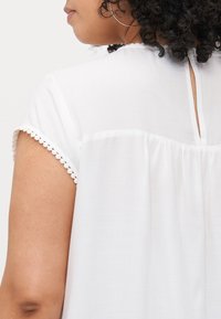 White blouse with short, scalloped sleeves and a keyhole back detail. Soft fabric, gathered back design, minimalistic and elegant.