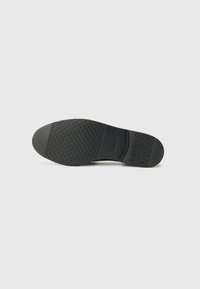 Semelle de chaussure en caoutchouc noir avec un motif texturé, présentant un design de vagues subtils près du bord et un logo de la marque en relief sur le côté.
