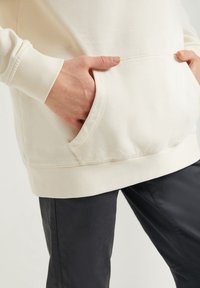 Světle béžová kapucí mikina s přední kapsou a žebrovan ými manžetami, kombinovaná s tmavými kalhotami, představující pohodlný a ležérní vzhled.