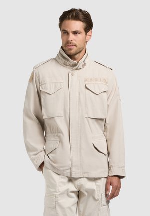 Man wearing beige utility jacket and matching cargo pants, standing with one hand in pocket, looking slightly to the side.