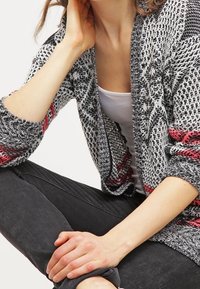 Stickad cardigan med ett mönster i svart, vitt och rött, öppen framtill, långa ärmar och textilstruktur. Bärs över en vit tanktop.