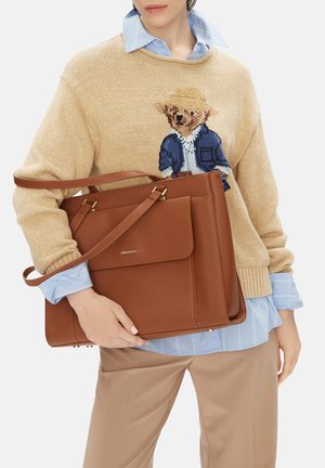 Persona que lleva un suéter beige con un gráfico de oso, camisa azul claro, pantalones beige, y sostiene un gran bolso de cuero marrón con correa para el hombro.