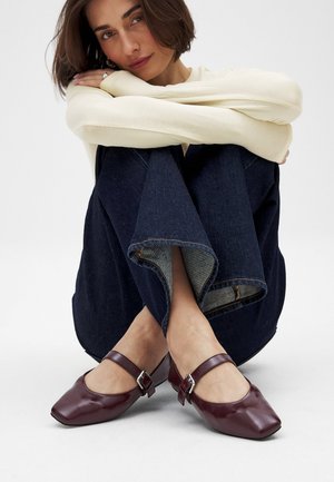 Bordeaux leren Mary Jane schoenen met vierkante neuzen en verstelbare enkelbandjes, geplaatst op een oppervlak met er bovenop gevouwen donkere spijkerbroeken.