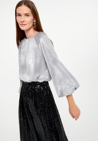 Glänzende silberne Bluse mit langen Ärmeln und gerafftem Ausschnitt, kombiniert mit einem plissierten schwarzen Paillettenrock, der einen strukturierten Kontrast schafft.
