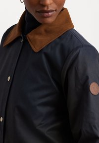 Navy jacket with a brown corduroy collar, featuring silver snap buttons and a round leather logo patch on the sleeve. Smooth texture.