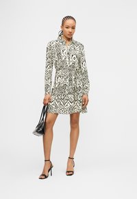 Γυναίκα με πλεγμένα μαλλιά, φορώντας μακρυμάνικο φόρεμα με ασπρόμαυρο μοτίβο, μαύρα πέδιλα με ψηλό τακούνι και κρατώντας μαύρη τσάντα χειρός.