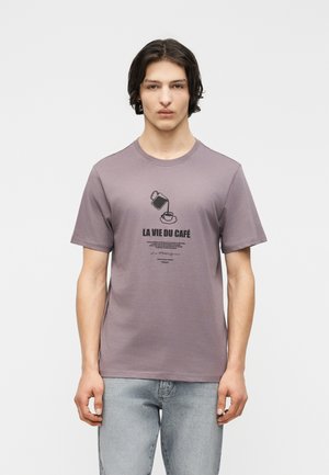 Νεαρός με σκούρα μαλλιά που φοράει μια απαλή μοβ μπλούζα με στάμπα καφετιέρα που ρίχνει καφέ σε φλιτζάνι και το κείμενο "LA VIE DU CAFÉ", συνδυασμένη με ανοιχτόχρωμο τζιν.