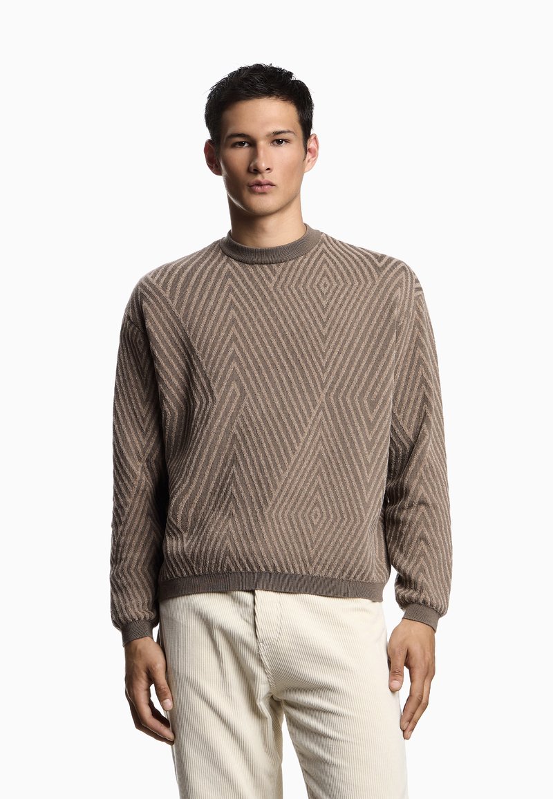 Maglione marrone a maglia con un motivo geometrico, collo rotondo e polsini a coste. Indossato sopra pantaloni in velluto a coste di colore chiaro.