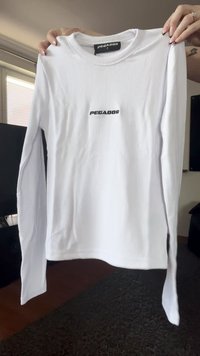 Camisa blanca de manga larga sostenida por manos, con el logo "PEGADOR" centrado en el pecho y la etiqueta de tamaño visible dentro del cuello.