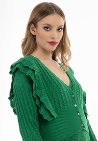 Turbulentsgrön stickad cardigan med volangärmar, v-ringning och fem dekorativa knappar. Texturerat mönster med öppna sömdetaljer.