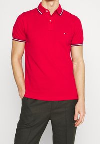 Rotes Poloshirt mit klassischem Kragen, kurzen Ärmeln und kontrastierenden schwarz-weißen Streifen am Kragen und an den Ärmeln. Kleines Logo auf der Brust.
