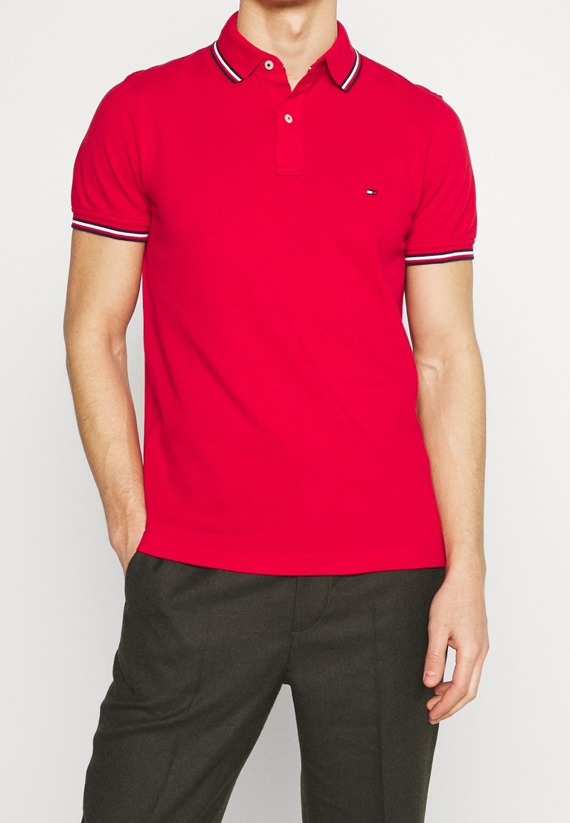 Rotes Poloshirt mit klassischem Kragen, kurzen Ärmeln und kontrastierenden schwarz-weißen Streifen am Kragen und an den Ärmeln. Kleines Logo auf der Brust.