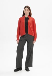 Rode geribbelde ritssportcardigan over een zwarte top, gecombineerd met grijze wijde broek en zwarte boots. Strakke lijnen, casual pasvorm, gestructureerde stof.