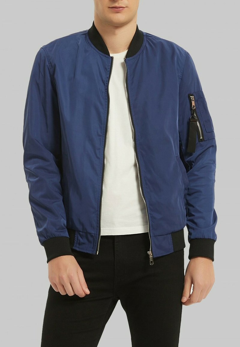 Veste bomber bleu marine en matériau léger, avec une fermeture éclair frontale, des poignets côtelés et une poche zippée sur le côté. Portée sur une chemise blanche.