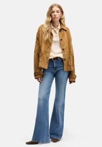 Beige suède jas met een kraag, knoopsluiting en twee steekzakken aan de voorkant, gedragen over een crèmekleurige trui en blauwe flare jeans. Bruine schoenen maken de look compleet.