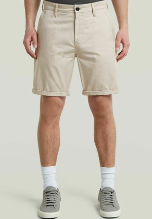 Homme portant un short beige retroussé, des chaussettes blanches montantes et des baskets grises, debout devant un fond clair uni.