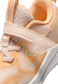 Zapatilla deportiva de color naranja claro con parte superior de malla, superposiciones texturizadas, cierre de cordones y una tira de velcro. Presenta acentos y logotipos en blanco.
