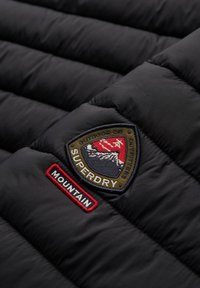 Tissu de veste matelassée noire avec un patch cousu montrant un logo de montagne et les mots "Superdry" et "Mountain".