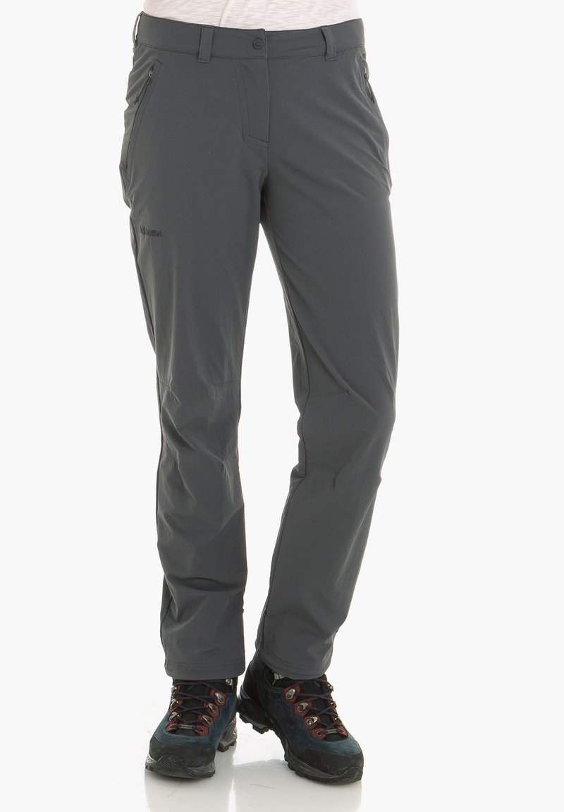 Pantalon d'extérieur gris foncé porté avec des chaussures de randonnée noires et rouges, visible sur une personne debout avec un haut blanc partiellement visible.