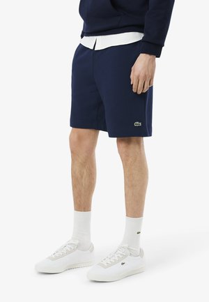 Shorts marine en mélange de coton, présentant une texture lisse et un détail de logo vert. Associés à des baskets blanches et des chaussettes à la cheville.