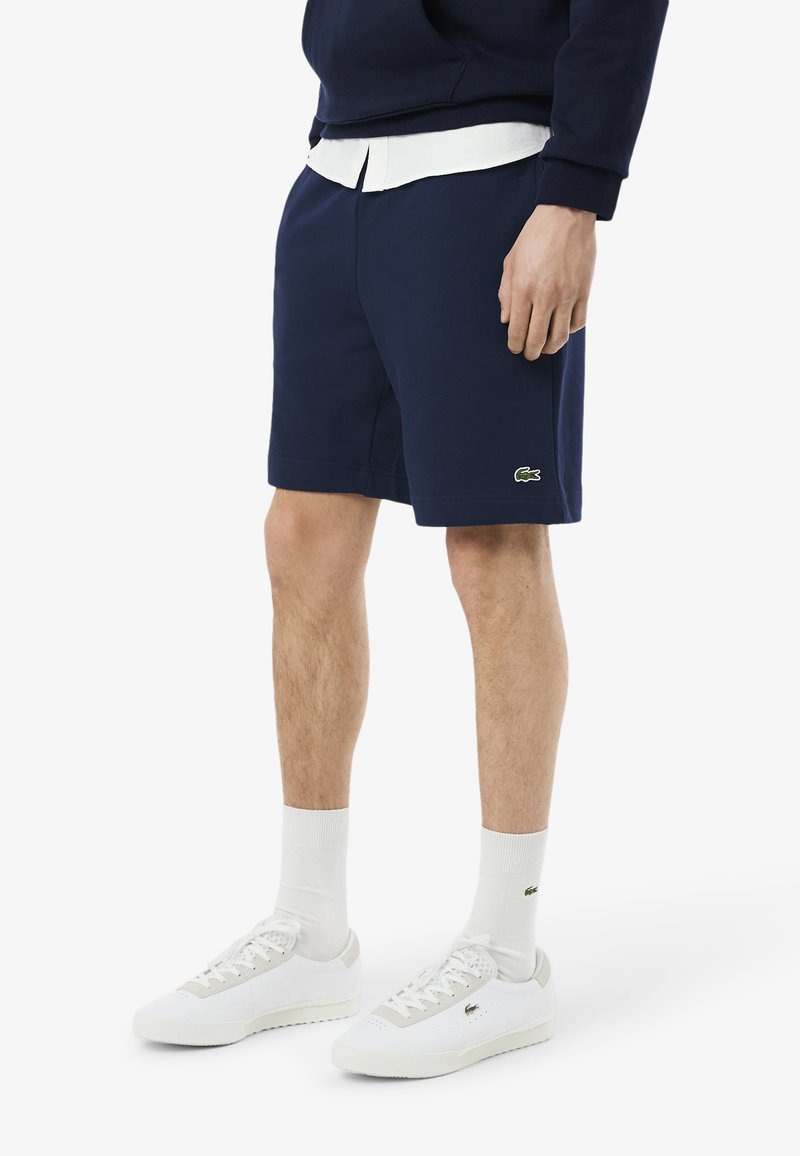 Shorts blu navy in misto cotone, con una texture liscia e dettagli del logo verdi. Abbinati a scarpe da ginnastica bianche e calzini alla caviglia.