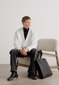 Bolso tote de cuero negro con asas planas, llevado por una persona que viste un cárdigan texturizado blanco sobre un cuello de tortuga negro y pantalones oscuros, sentada en una silla clara.