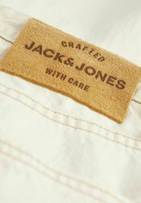 Smeđa plišana etiketa s utisnutim tekstom "CRAFTED JACK & JONES WITH CARE" na krem tkanini, s kontrastnim šavovima duž ruba.