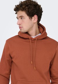 Roestkleurige hoodie met een voorzak, capuchon met trekkoord en gerafelde stof. Voorzien van trekkoorden met bijpassende uiteinden.