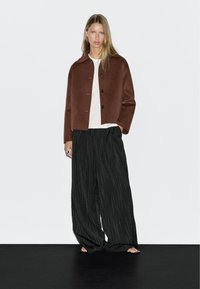 Giacca corta in misto lana marrone con colletto tondo e quattro bottoni, abbinata a pantaloni a gamba larga neri a righe per la vita alta.