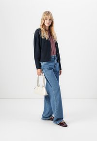 Jeune femme portant un cardigan marine, un chemisier mauve, un jean large bleu et des chaussures bordeaux, tenant un petit sac à main blanc devant un fond blanc.