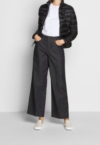 Sort pufferjakke, hvid t-shirt, højtsiddende mørk denim vide bukser og cremede slip-on sko. Har synlige sømme og lommer.
