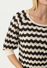 Pull en tricot noir et crème avec un motif ondulé, manches courtes bouffantes et encolure côtelée, mettant en avant une texture ajourée et des couleurs contrastées.