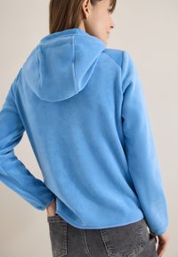 Femme portant un sweat à capuche en polaire bleu clair, capuche relevée, debout, main dans la poche arrière d'un jean gris foncé, sur un fond uni.