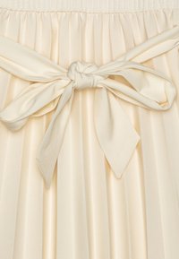 Pehmeäbeige satiinista valmistettu laskostettu hame, jossa on teksturoitu vyötärönauha ja solmittava rusetti vyötäröllä, esittäen sileää ja virtaavaa kangasta.