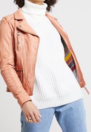 Personne portant une veste en cuir marron clair par-dessus un pull à col roulé en tricot blanc et un jean en denim bleu clair.