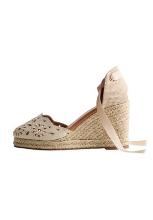 Alpargata cuña beige con tacón de yute trenzado, patrones florales recortados en la puntera y lazos para anudar en el tobillo sobre fondo blanco.