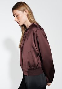 Giacca bomber bordeaux in satin liscio con bordo a coste, tasche laterali e colletto rialzato. La vista laterale mette in evidenza il design slim fit.