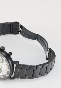 Montre en métal noir avec un bracelet à maillons, texture lisse, un cadran rond avec des chiffres romains et un mécanisme de fermoir.