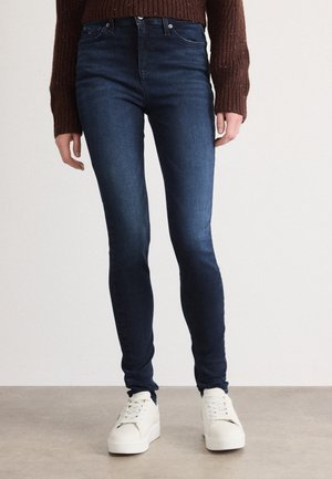 Person wearing dark blue skinny jeans, brown knitted sweater, and white sneakers standing on a neutral floor with a plain background.