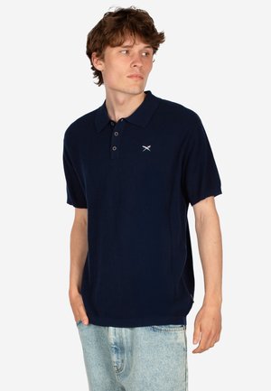 Junger Mann, der ein marineblaues Polohemd mit kurzen Ärmeln und kleinem Logo trägt, kombiniert mit hellblauen Jeans, schaut zur Seite.