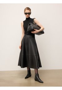 Nais, kes kannab musta ilma varrukateta kõrge kaelusega pluusi, nahast midi seelikut, pahkluu saapaid ja päikeseprille, hoides suurt nahast käekotti ühtlase tausta ees.