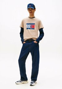 Ung mand iført en beige Tommy Jeans T-shirt over en navy langærmet skjorte, mørkeblå jeans, hvide sneakers og en blå kasket, stående med hænderne i lommen.