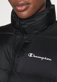 Svart pufferjacka med hög krage, försedd med dragkedjestängning och en vit Champion-logotyp. Materialet är slätt med en lätt lyster.