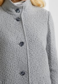 Grå ullkappa med en texturerad yta, hög krage och fyra stora, mörka knappar fram, vilket betonar en strukturerad design.