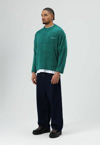 Groene fleece trui met een gestructureerde afwerking, gecombineerd met losse donkere spijkerbroeken en zwarte schoenen met roze veters.