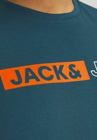 Teal bomulds T-shirt med et stort orange rektangulært grafisk design og sort tekst "JACK&" samt et mindre hvidt logo til højre.