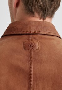 Braune Lederjacke mit sichtbarem Kragen, deutlich erkennbaren Nähten und einem kleinen Stern-Emblem auf dem Rücken nahe dem Halsausschnitt.