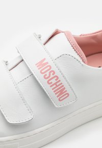 Zapatilla blanca de cuero con detalles en rosa y una correa de Velcro. El nombre de la marca "MOSCHINO" se muestra de manera prominente en rosa en la correa.