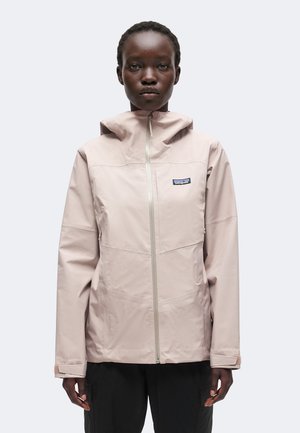 Persona joven con una chaqueta con capucha beige claro con cremallera y un pequeño logo de Patagonia en el pecho, de pie frente a un fondo sencillo.