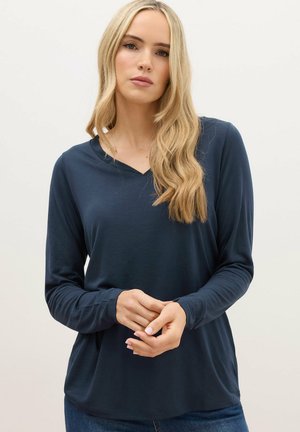 Blonde Frau mit lockeren Wellen, die ein marineblaues langärmliges Shirt mit V-Ausschnitt trägt und vor einem schlichten hellen Hintergrund steht.